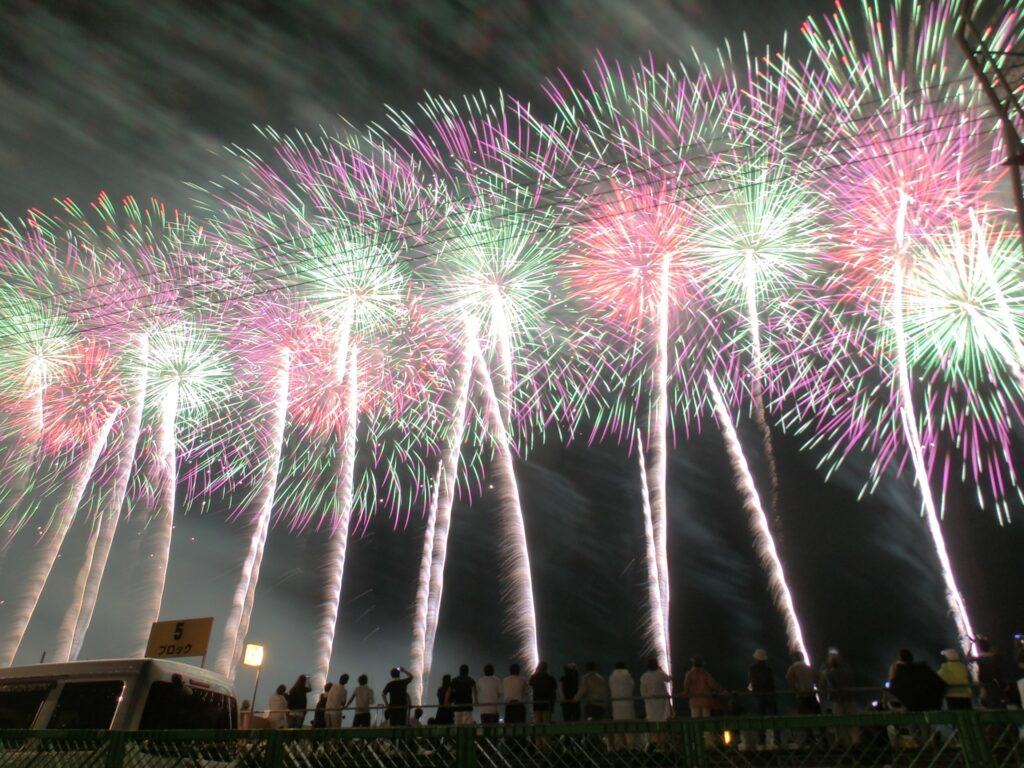 大曲花火大会で、観客の頭上に広がる緑と紫の大輪花火が夜空を埋め尽くす様子